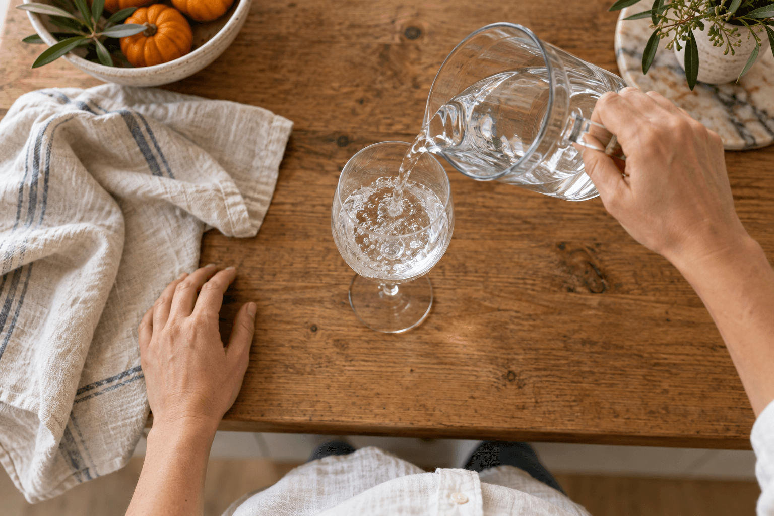 Overhead view of adult hands pouring water from a clear glass pitcher into a stemmed glass on a wooden kitchen surface.