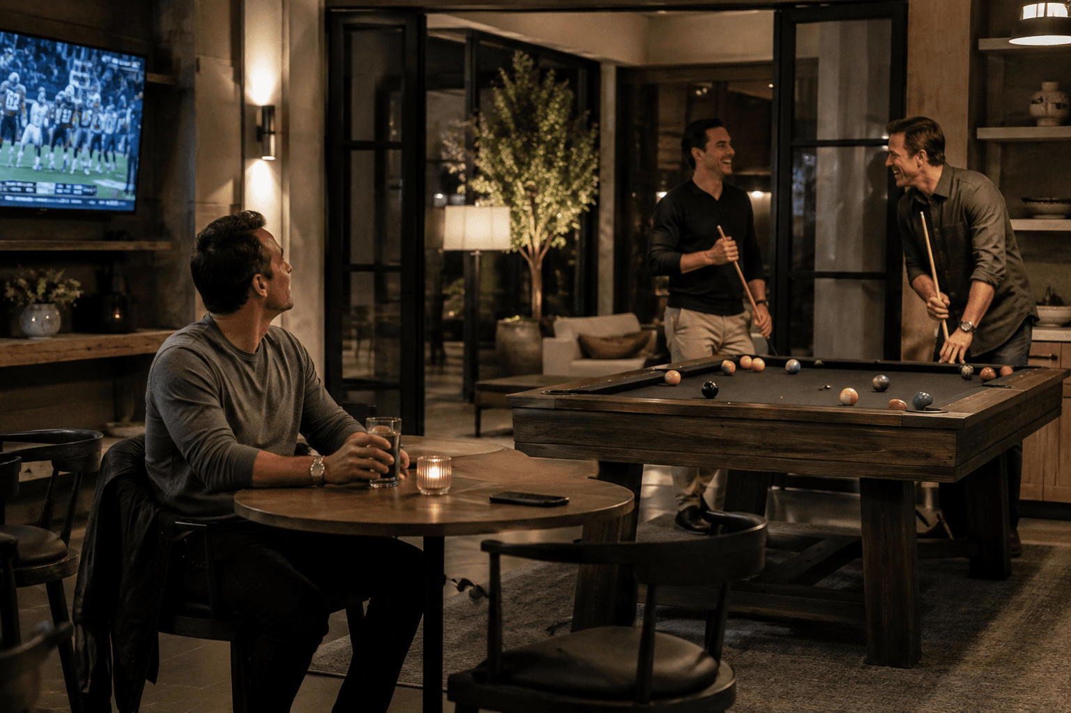 Three longtime male friends in a modern game lounge, with two laughing around a pool table while another sits nearby watching the television.